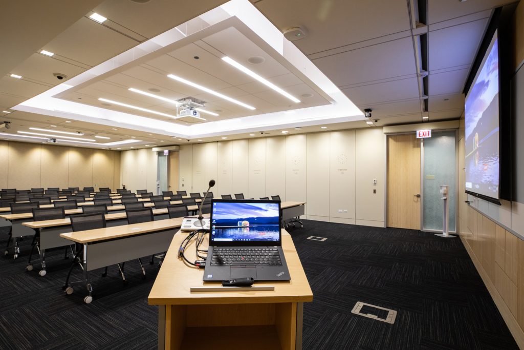Conference Room 1AB at One Rotary Place. Photo of a large conference room with a podium &amp; microphone,projector, and screen.