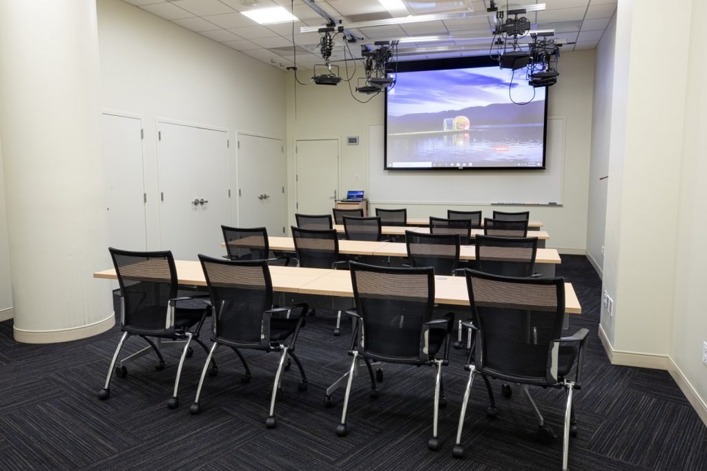 1st Floor Studio Conference Room at One Rotary Place. Photo of a small conference room with tables &amp; chairs, a projector, and screen.