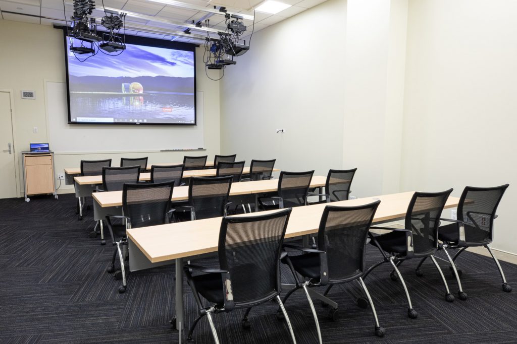 1st Floor Studio Conference Room at One Rotary Place. Photo of a small conference room with tables &amp; chairs, a projector, and screen.