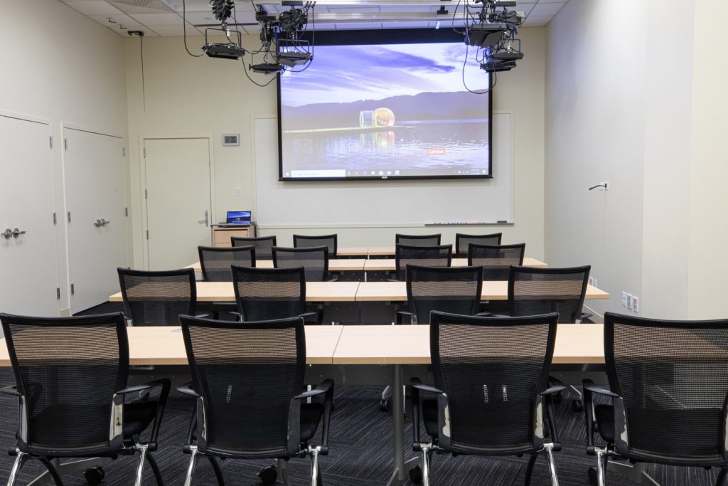 1st Floor Studio Conference Room at One Rotary Place. Photo of a small conference room with tables &amp; chairs, a projector, and screen.