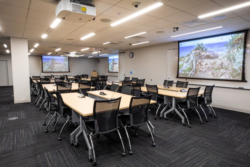Conference Room 3AB at One Rotary Place. Photo of a large conference room with tables &amp; chairs, microphones, and multiple projectors &amp; screens.