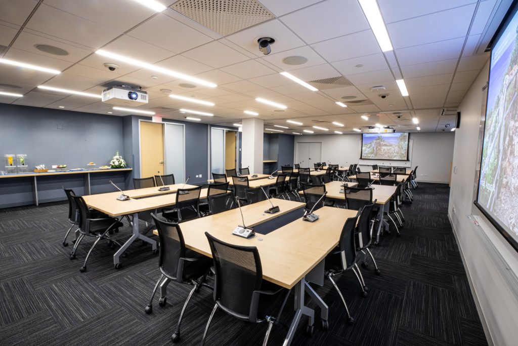 Conference Room 3AB at One Rotary Place. Photo of a large conference room with tables &amp; chairs, microphones, and multiple projectors &amp; screens.