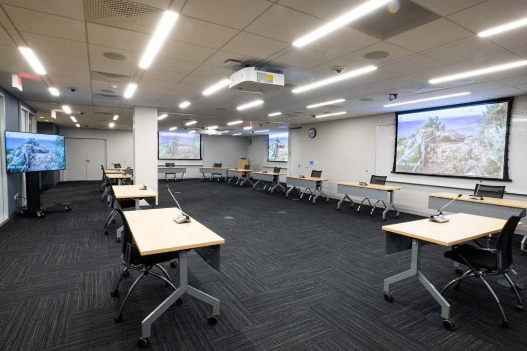 Conference Room 3AB at One Rotary Place. Photo of a large conference room with tables &amp; chairs, microphones, and multiple projectors &amp; screens.