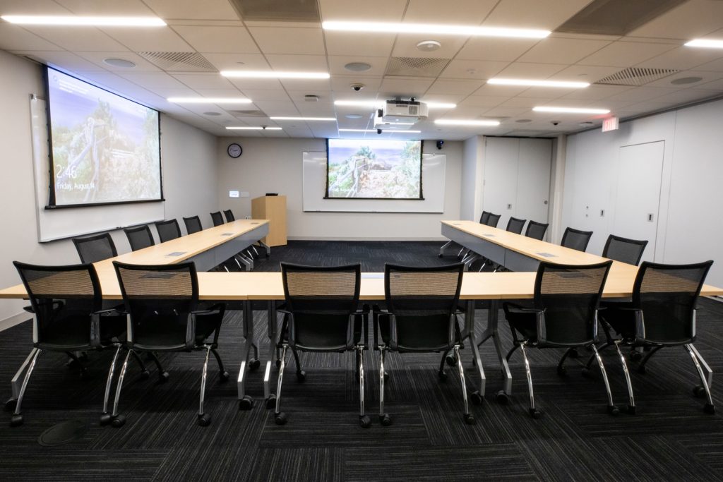 Conference Room 3B at One Rotary Place. Photo of a large conference room with tables &amp; chairs, a podium, multiple projectors &amp; screens, and multiple whiteboards.