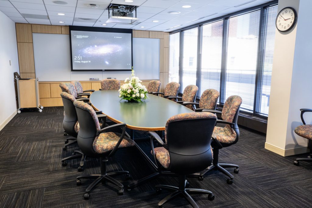 3 East Conference Room at One Rotary Place. Photo of a an executive conference room with a center table, projector &amp; screen, and a whiteboard.