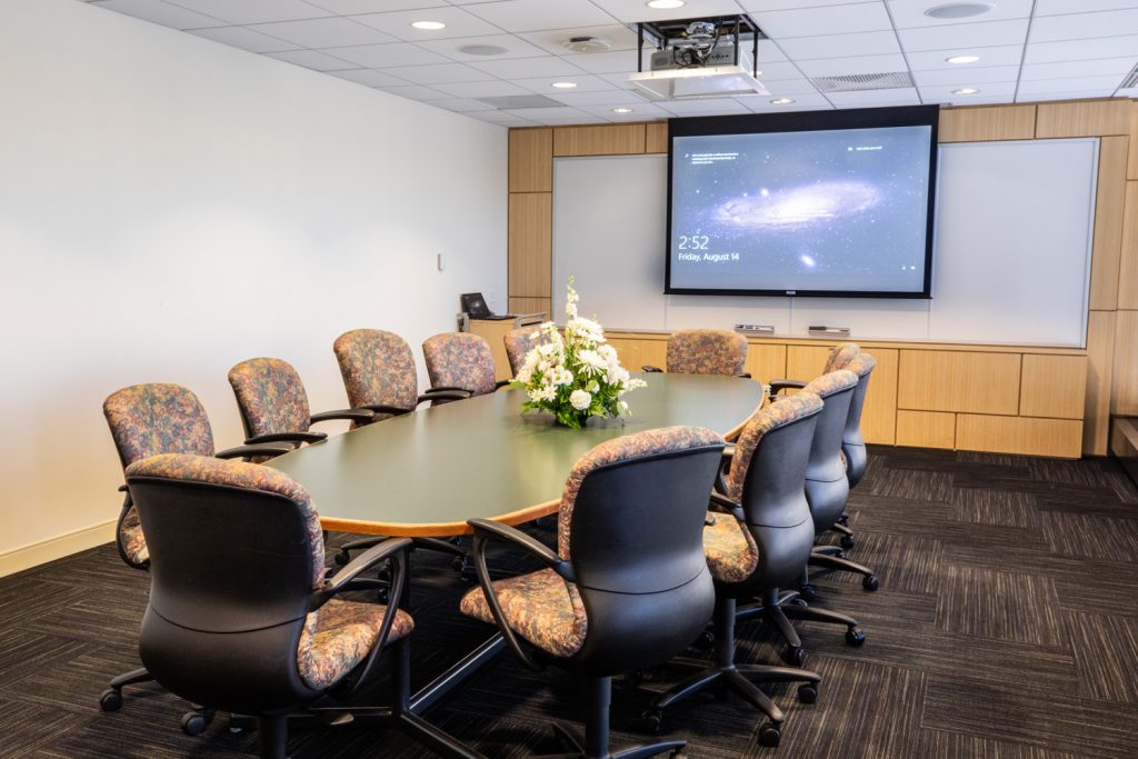 3 East Conference Room at One Rotary Place. Photo of a an executive conference room with a center table, projector &amp; screen, and a whiteboard.
